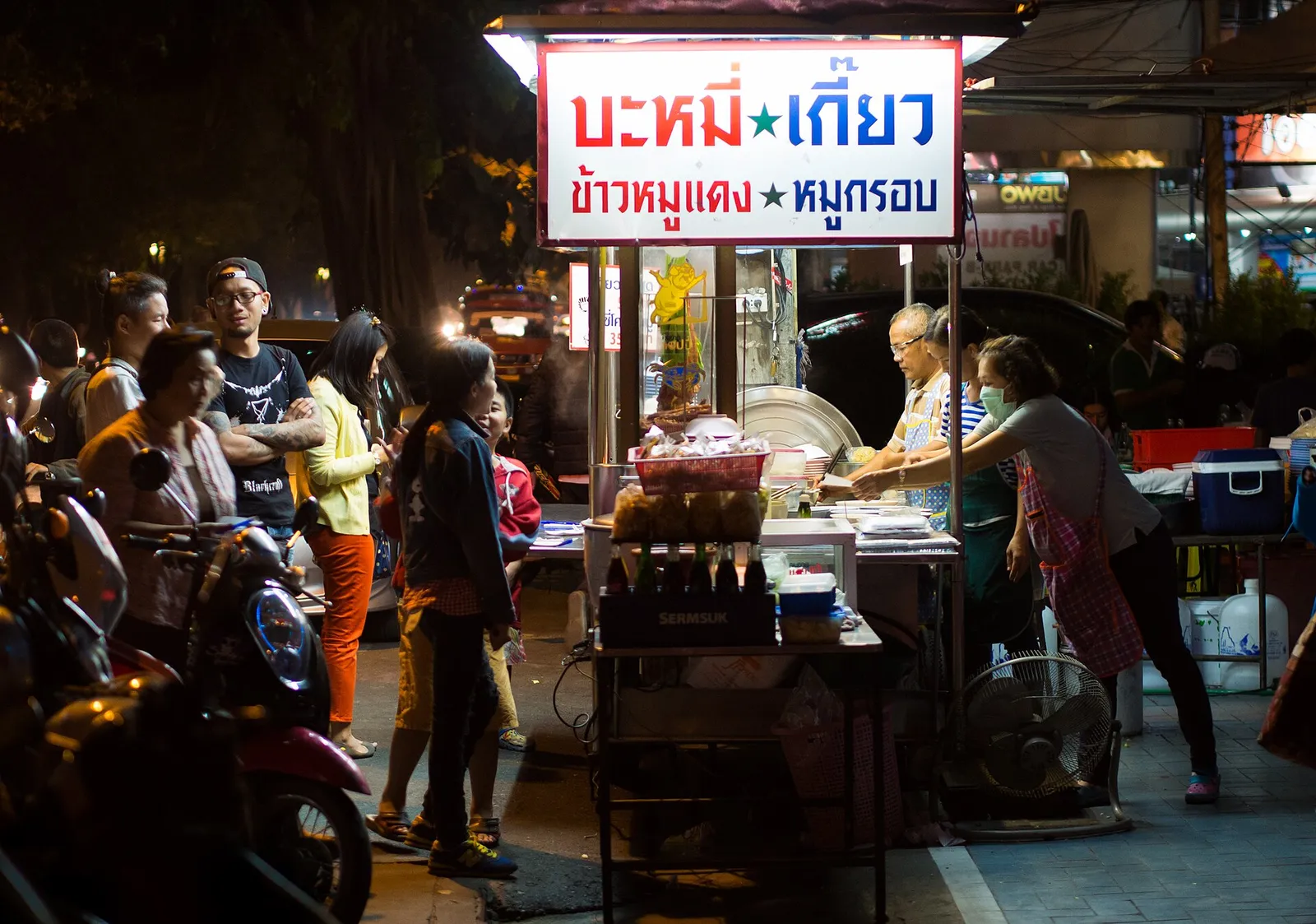 דוכן אטריות רחוב בבנגקוק תאילנד בלילה עם שלט תפריט תאי ולקוחות מזמינים אוכל